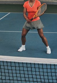 Tennispelaaja oranssissa lyhythihaisessa paidassa ja harmaissa hameessa, pitäen mailaa, sinisessä kentässä, jossa verkko on etualalla.
