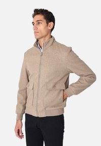 Giacca beige con zip realizzata in misto lana, con colletto alto, tasche laterali e polsini a costine. Texture liscia con taglio classico.