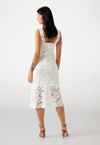 White lace dress featuring intricate floral patterns, fitted bodice, square neckline, and knee-length hem. Paired with strappy heels.