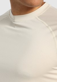 Maglietta sportiva beige chiaro a maniche corte con superficie texturizzata, dotata di scollatura tonda e dettagli di cucitura a contrasto.