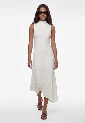 Mujer con vestido midi blanco texturizado sin mangas y cuello alto, gafas de sol oscuras y sandalias marrones con tiras, caminando hacia adelante sobre fondo blanco.