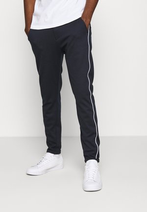 Uomo che indossa pantaloni della tuta blu scuro con piping bianco laterale e sneakers bianche, in piedi con le mani in tasca su uno sfondo semplice.