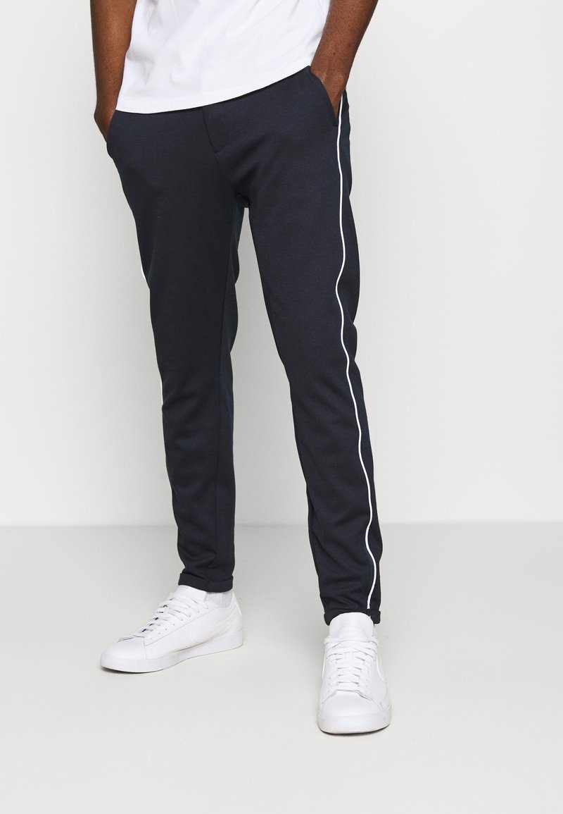 Homme portant un jogger bleu marine foncé avec un liseré blanc sur les côtés et des baskets blanches, debout les mains dans les poches sur un fond uni.