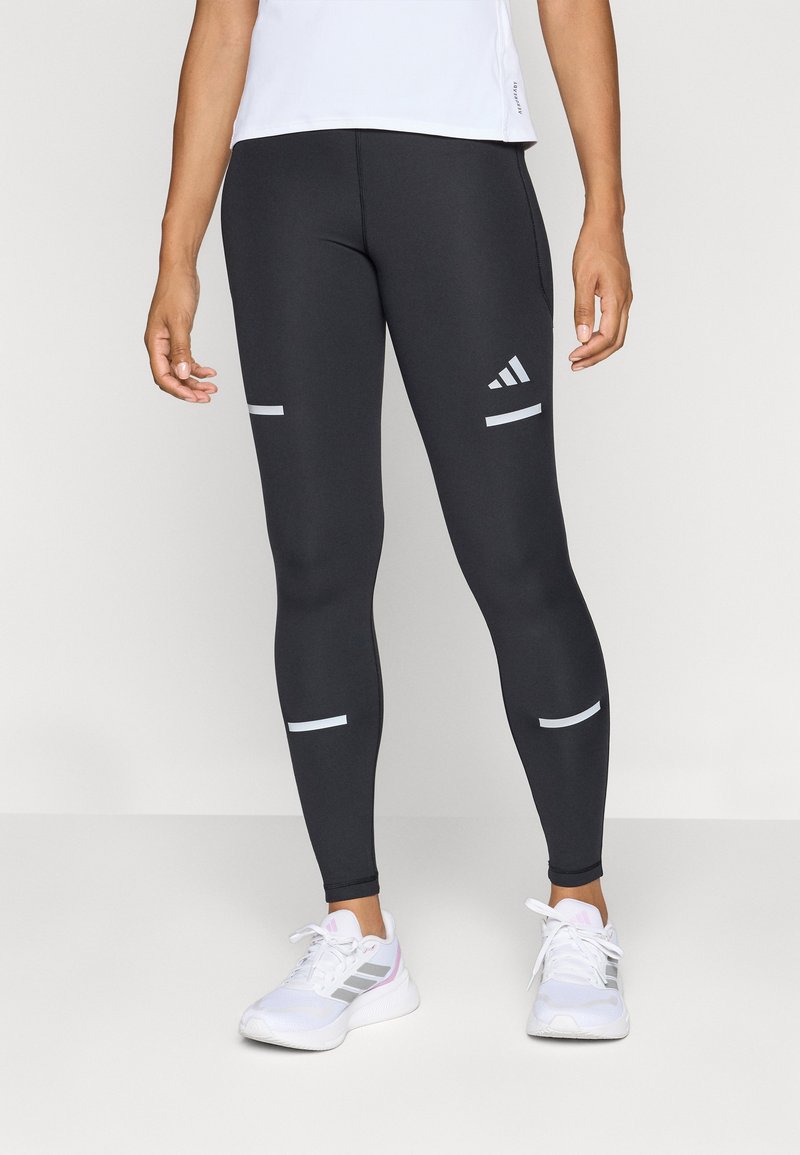 Leggings sportivi neri con accenti riflettenti argento, caratterizzati da una vestibilità aderente e una texture liscia. Abbinati a sneakers bianche.