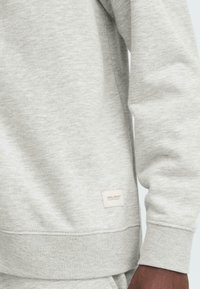 Light gray sweatshirt made of soft fabric. Features a ribbed hem and cuffs. A small brand label is stitched on the bottom left side.