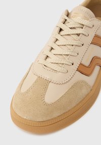 Beiger Sneaker mit Wildlederkappe, gewebten Schnürsenkeln, Laufsohle aus Kaugummigummi und braunem Logostreifen an der Seite.