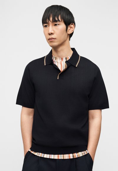 Άνδρας που φοράει μαύρο πόλο κοντομάνικο πάνω από πολύχρωμο ριγέ πουκάμισο, με τα χέρια στις τσέπες, κοιτώντας ελαφρώς πλάγια σε απλό φόντο.