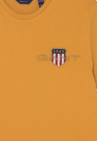 Żółta bawełniana koszulka z okrągłym dekoltem i haftowanym logo w kształcie tarczy z czerwonymi i niebieskimi paskami. Napis brzmi "GANT 1949."