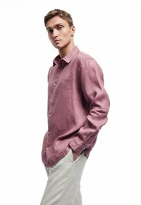 Camisa de lino rosa claro con mangas largas, delantero abotonado y corte casual, combinada con pantalones de textura en un color claro.
