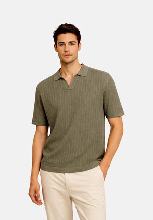 Mann trägt olivgrünes, kurzärmeliges, gestricktes Polohemd und hellbeige Hose, steht mit einer Hand in der Tasche vor neutralem Hintergrund.