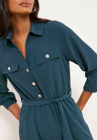 Türkisfarbenes Hemdkleid mit Kragen, zwei Brusttaschen und langen Ärmeln. Verfügt über eine taillierte Passform mit Gürtel und einen weichen, glatten Stoff. Goldene Knöpfe verleihen dem Detail zusätzlichen Charme.