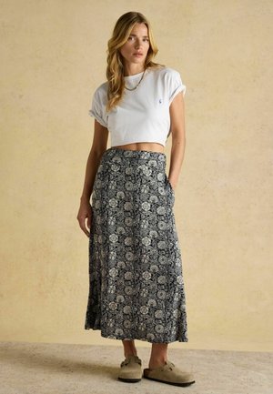 Naine, kellel on seljas valge lühike T-särk, must-valge lillemustriga maxi-seelik, beežid libisemisjalatsid ja kuldne kaelakee, seisab beežil põrandal.