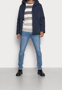 Mörkblå pufferjacka med huva, bärs över en randig stickad tröja. Ljust blå jeans och svarta ankelstövlar fulländar outfiten.