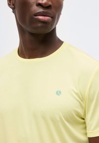 Κοντινό πλάνο ενός ατόμου με σκούρο δέρμα που φοράει ένα απαλό κίτρινο υφασμάτινο t-shirt με στρογγυλή λαιμόκοψη και μικρό ανοιχτό μπλε λογότυπο στο στήθος.