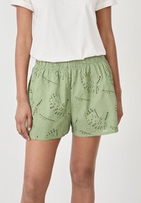 Shorts verts brodés avec un motif floral, comportant des découpes en œillet et une taille élastique. Portés avec un t-shirt blanc uni.