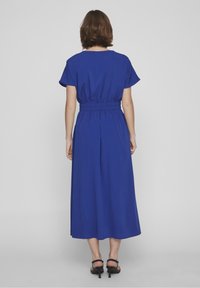 Modré šaty ke kolenům s krátkými rukávy, nařaseným pasem a splývavou sukni. Hladká textura s ozdobným prvkem na zádech. Černé sandály na podpatku.