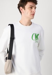 Fehér pamut pulóver zöld "CK" logóval; kerek nyakkivágással és bordás mandzsettával rendelkezik. Szürke táskával és fekete pánttal viselve.