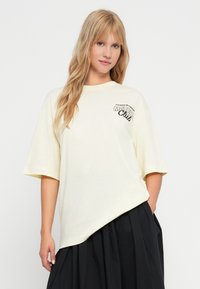 Lys gul oversized t-skjorte med korte ermer og rund hals. Har et trykt grafisk motiv plassert på venstre bryst.