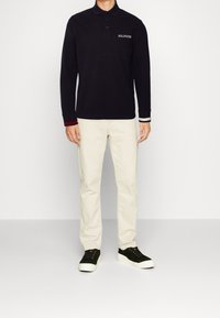 Mörkblå pikétröja med lång ärm, tre-knappara knäppning och "HILFIGER" logotyp. Kombineras med ljusbeiga byxor och mörkgröna sneakers.