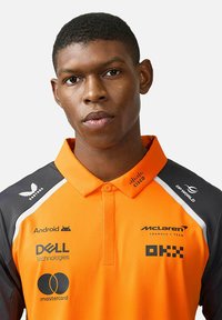 Pomarańczowa koszulka polo z kołnierzykiem, z szarymi rękawami oraz logotypami McLaren, Dell i innych sponsorów. Teksturowana tkanina z cienkimi paskami.