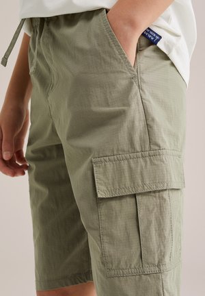 Gyermek világos olívazöld cargo rövidnadrágban, oldalsó zsebbel, kezét az első zsebben tartva, fehér pólóval párosítva.