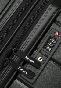 Valigia rigida nera con doppie cerniere, maniglie firmate e lucchetto a combinazione approvato dalla TSA. Superficie texturizzata e bordi rinforzati visibili.