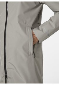 Chaqueta impermeable de color gris claro con cremallera frontal, bolsillos laterales y una textura suave. Incluye un acento reflectante en la manga.