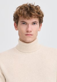 Jonge man met krullend lichtbruin haar, blauwe ogen, draagt een beige geribbelde coltrui, kijkt recht vooruit tegen een effen witte achtergrond.