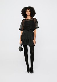 Schwarzes halbtransparentes Oberteil mit kurzen Ärmeln, kombiniert mit einem schwarzen plissierten Minirock und schwarzen Strumpfhosen, ergänzt durch spitze schwarze High Heels.