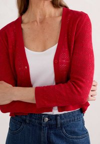 Roter Strickcardigan mit strukturiertem Muster, cropped Design, Knopfverschluss und langen Ärmeln, getragen über einem weißen Oberteil.