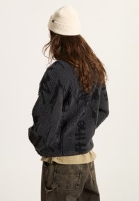 Schwarze Fleecejacke aus strukturiertem Material, mit lockerer Passform und gerippten Bündchen, über einem beigen Hemd getragen, kombiniert mit dunklen Hosen.