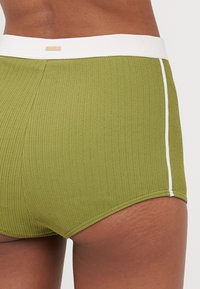 Olivengrønne svømshorts med ribbet overflade, hvid elastisk talje og sideliste. Stoffinishen er tekstureret, og designet er tætsiddende.
