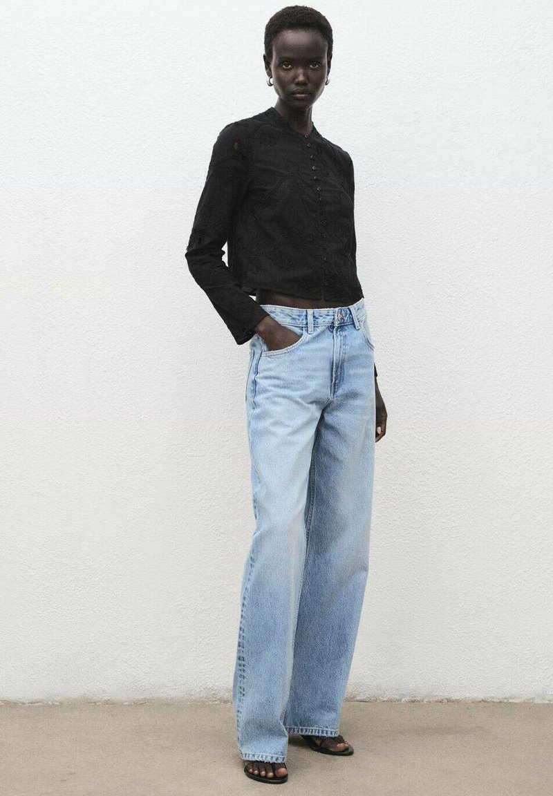Camisa negra corta de botones con tejido texturizado, combinada con pantalones de mezclilla de pierna ancha de color azul claro. Modelo vistiendo sandalias negras, de pie contra un fondo neutral.