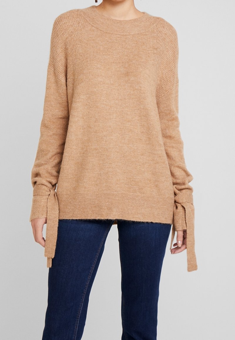Femme portant un pull beige à manches longues avec des détails noués sur les côtés et un jean bleu foncé, sur un fond gris uni.