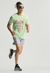 Camiseta deportiva de color verde claro con un diseño gráfico rosa y el texto "BERLÍN 2025", combinada con pantalones cortos grises y zapatillas de running negras.