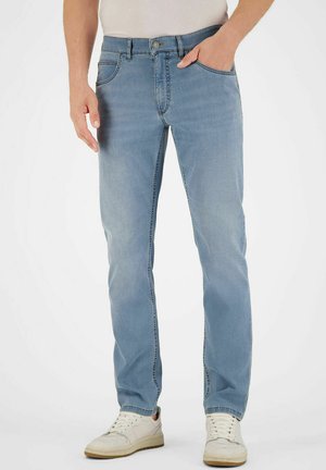 Mann trägt hellblaue Jeans und weiße Turnschuhe, steht mit einer Hand in der Hosentasche vor einem einfarbigen Hintergrund.