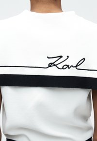 Οπίσθια όψη ενός ατόμου που φοράει λευκό μπλουζάκι με μαύρη οριζόντια γραμμή και τη λέξη "Karl" κεντημένη με μαύρα καλλιγραφικά γράμματα πάνω από τη γραμμή.