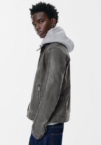 Giovane uomo che indossa una giacca di pelle grigia sopra un hoodie grigio chiaro e jeans blu, con uno sguardo laterale e un'espressione neutra.