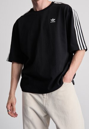 Person trägt ein schwarzes Adidas-T-Shirt mit weißen Streifen an den Ärmeln und hellbeigen Hosen, steht mit einer Hand in der Tasche.