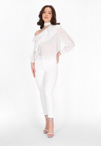 Weiße schulterfreie Bluse mit schwarzen Punkten, Rüschenausschnitt und langen Ärmeln, kombiniert mit weißen, taillierten Jeans und silbernen, geschnürten High Heels.