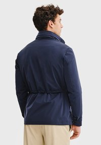 Granatowa wodoodporna kurtka z podwyższonym kołnierzem, elastycznym sznurkiem w talii oraz zapinanymi mankietami. Gładka tekstura, elegancki design.