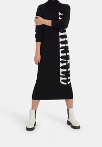 Svart stickad klänning med långa ärmar och hög krage, med en djärv vit textdesign på ena sidan. Bärs med vita ankelskor.