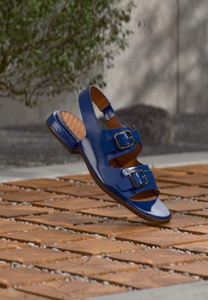 Sandale bleue brillante avec deux boucles, sangle arrière, flottant au-dessus d'une surface en carreaux de bois avec un fond de plante verte et de pierre.