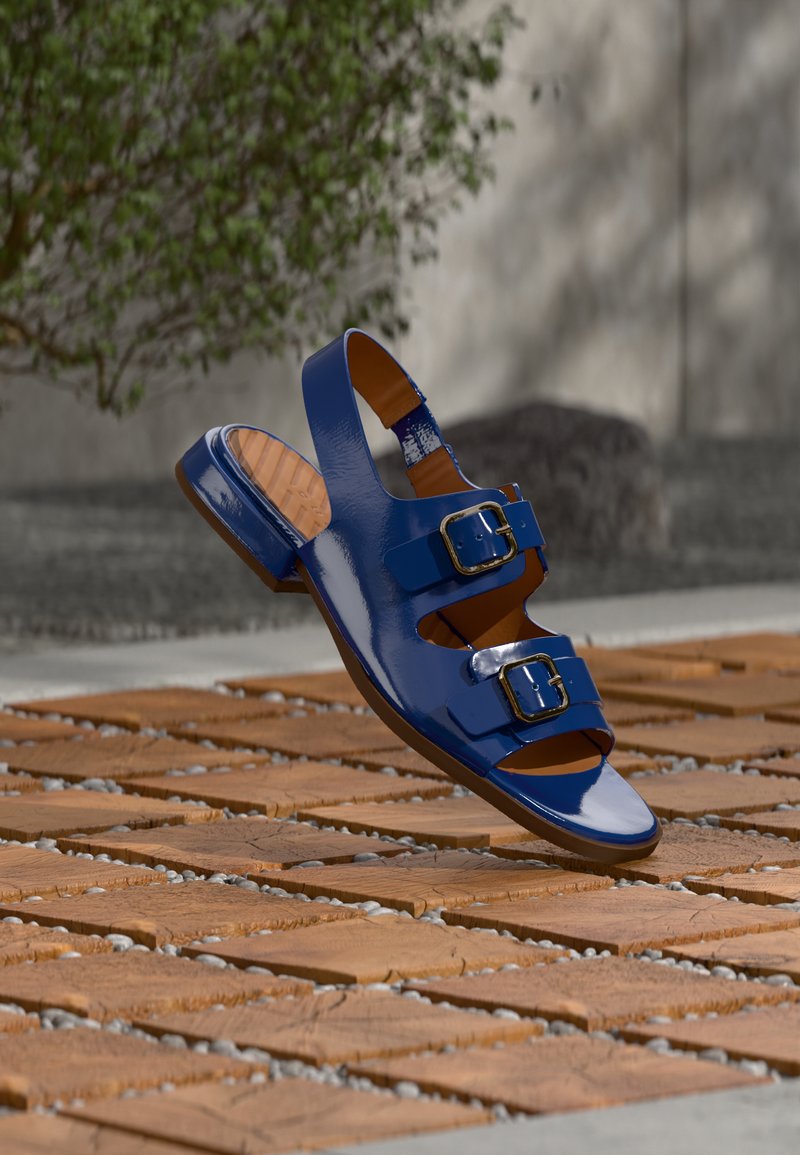 Sandale bleue brillante avec deux boucles, sangle arrière, flottant au-dessus d'une surface en carreaux de bois avec un fond de plante verte et de pierre.