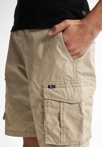 Pantaloni cargo beige realizzati in tessuto leggero, dotati di tasche laterali e una piccola etichetta del marchio. Superficie testurizzata e vestibilità casual.