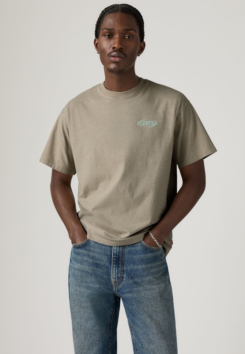 Jeune homme portant un t-shirt Levi's marron clair et un jean bleu, debout les mains dans les poches devant un fond uni.