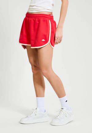 Mujer con pantalones cortos deportivos rojos con ribete blanco, zapatos deportivos blancos y calcetines blancos de caña media, posando contra un fondo liso.