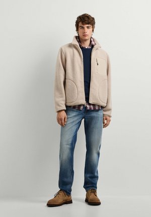 Giovane uomo che indossa una giacca in pile beige sopra un maglione blu navy e una camicia a quadri, jeans blu e scarpe marroni con lacci, in piedi davanti a un muro semplice.