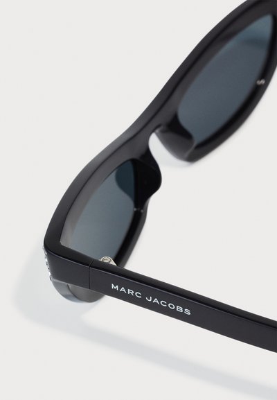 Lunettes de soleil noires avec une monture solide, des verres foncés et une marque en argent sur le côté. Elles présentent un design rectangulaire et une texture lisse.