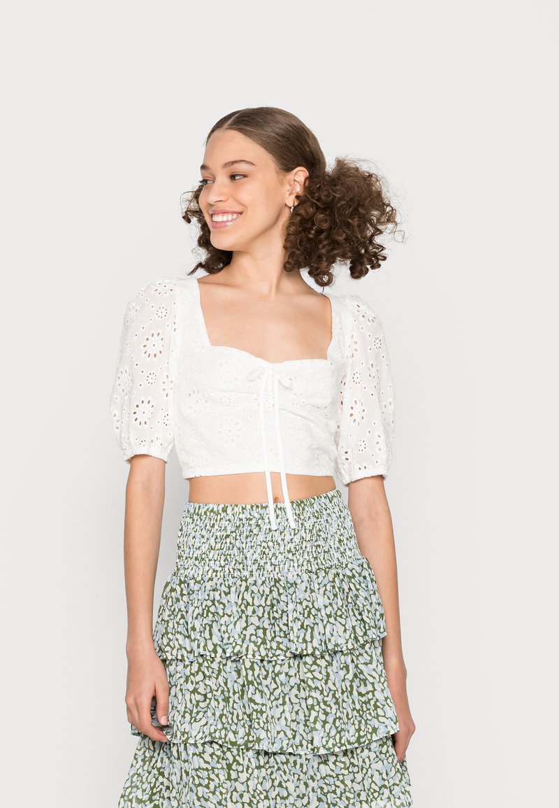 Top cropped branco com mangas bufantes, design de ilhós florais e detalhe de cordão, combinado com uma saia rodada de camadas em verde e azul claro.